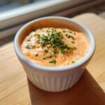 A close-up of creamy, orange-tinted remoulade sauce topped with fresh parsley and diced onions in a white bowl.
