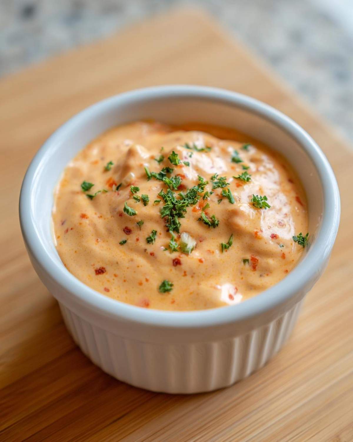 A close-up of creamy, orange-hued remoulade sauce garnished with fresh parsley in a white ramekin.