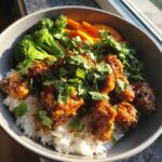 Close-up of a bowl containing white rice topped with saucy chicken, steamed broccoli, carrots, and fresh cilantro.