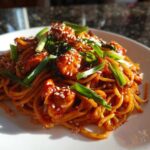 A close-up of 20-Minute Saucy Gochujang Chicken Noodles tossed in a bright red sauce, topped with green onions and sesame seeds.