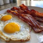 Close-up of two sunny-side-up eggs next to crispy strips of Air Fryer Bacon with Eggs on a white plate.
