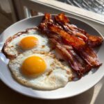 Two sunny-side-up eggs next to crispy strips of Air Fryer Bacon with Eggs on a white plate.