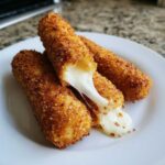 Close-up of crispy Air Fryer Mozzarella Sticks on a white plate, showing a dramatic cheese pull from one broken stick.