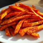 A close-up of bright orange Air Fryer Sweet Potato Fries coated heavily in red Cajun spice mix on a white plate.