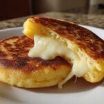 Two golden-brown Arepas Con Queso (Cornmeal Pockets) on a white plate, one split open revealing melted white cheese stretching out.