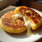 Two golden-brown Arepas Con Queso on a white plate, one split open revealing melted, stretching white cheese.
