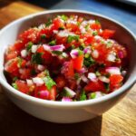 Close-up of a white bowl filled with vibrant Authentic Pico de Gallo, showing diced tomatoes, red onion, and cilantro.
