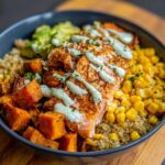A vibrant bowl featuring seasoned salmon, roasted sweet potato cubes, corn, quinoa, and avocado, drizzled with a light green sauce.