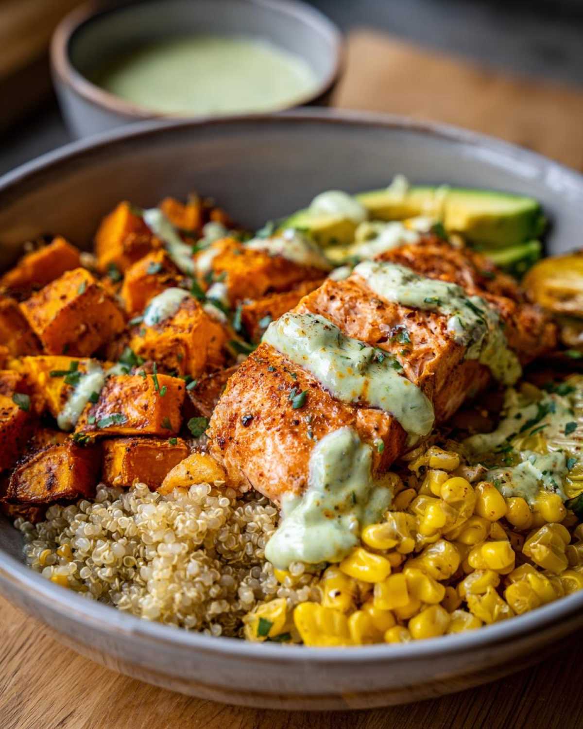 A close-up of a vibrant BBQ Salmon Bowls with Sweet Potato, featuring seasoned salmon, roasted sweet potato cubes, quinoa, corn, and avocado, drizzled with a green sauce.