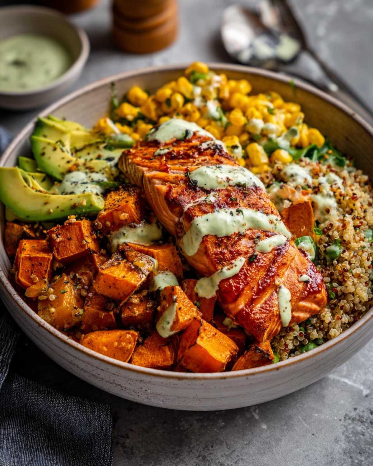 Close-up of a BBQ Salmon Bowl featuring grilled salmon, roasted sweet potato cubes, corn, and avocado, drizzled with green sauce.