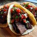 Three freshly made Carne Asada Tacos topped generously with vibrant pico de gallo salsa on a wooden cutting board.