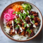 Close-up of a bowl featuring sticky glazed chicken over rice, topped with pickled onions, carrots, and mint, characteristic of Chicken Banh Mi Bowls.