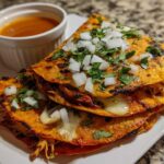 Three stacked, cheesy Chicken Birria Tacos topped with diced white onion and cilantro, served with a side of consomé for dipping.