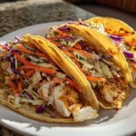 Three delicious Cod Fish Tacos loaded with flaky seasoned fish and vibrant crunchy slaw, served on a white plate.