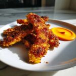 Close-up of several golden brown, crispy corn ribs served on a white plate next to a swirl of bright orange chili lime mayo.