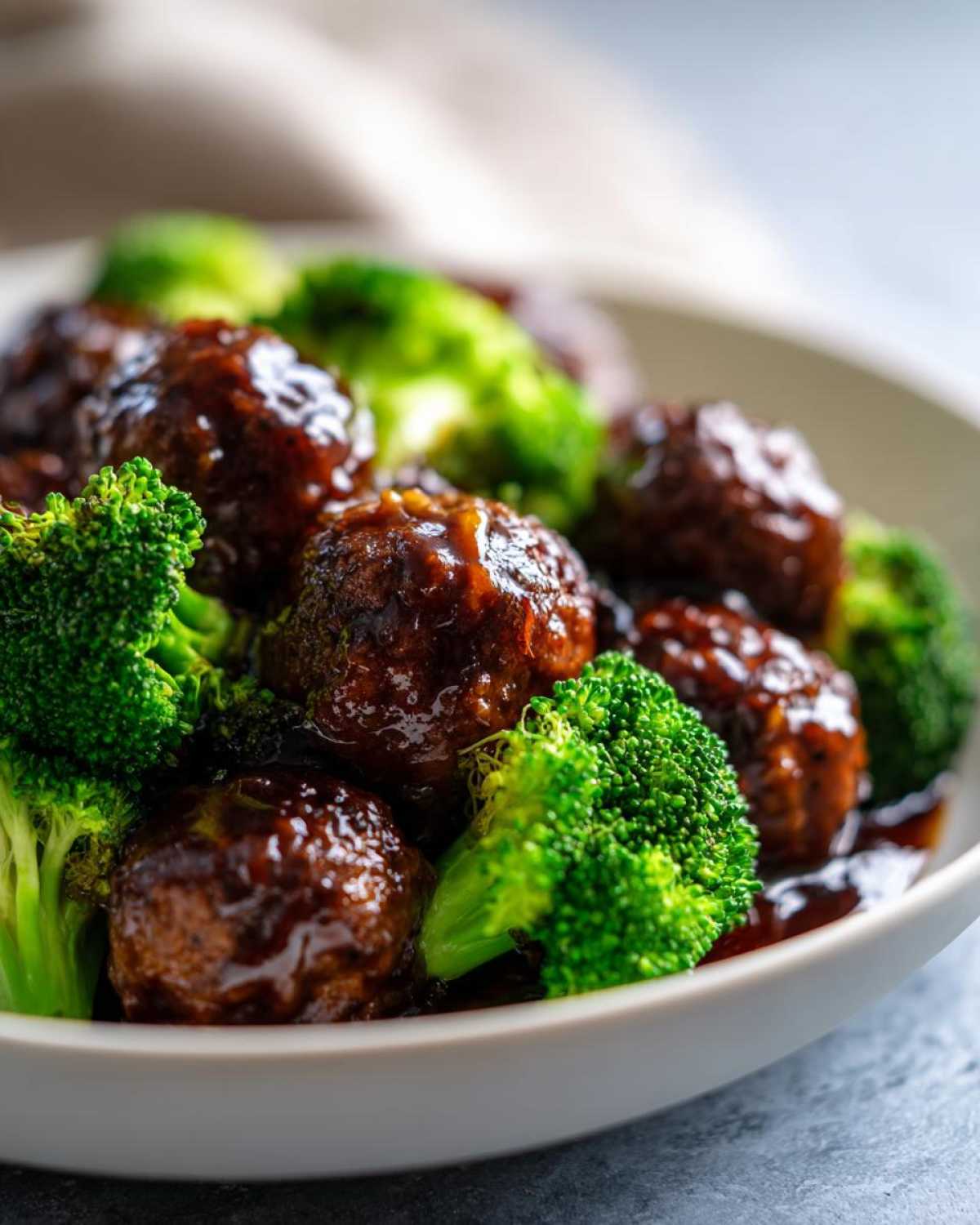 Close-up of glossy Crockpot Teriyaki Meatballs with bright green broccoli florets in a white bowl.