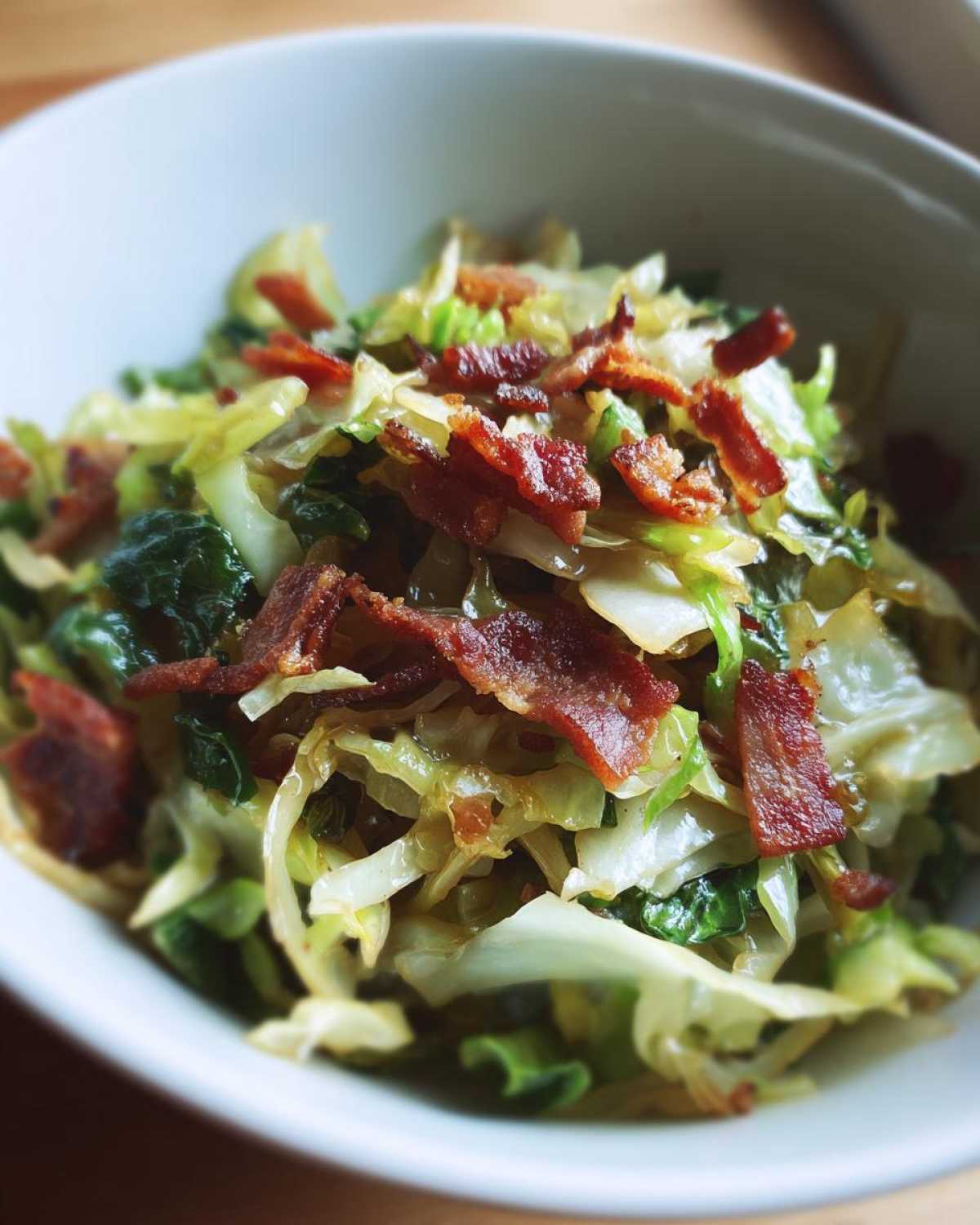 Close-up of a white bowl filled with tender Fried Cabbage with Crispy Bacon pieces scattered on top.