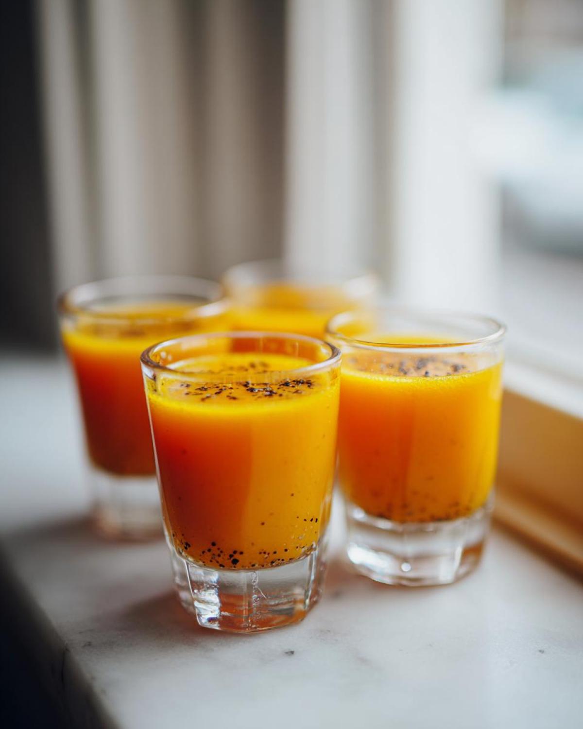 Four small glasses filled with bright orange Ginger Lemon Turmeric Shot, topped with black pepper.