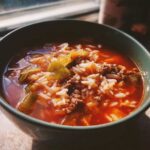 Close-up of a dark green bowl filled with steaming Golumpki Soup, featuring ground meat, white rice, and cabbage in a rich tomato broth.
