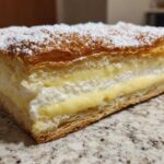 Close-up of a thick slice of Karpatka Cake (Polish Cream Cake) showing flaky pastry layers and rich vanilla cream filling, dusted with powdered sugar.