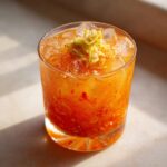 A vibrant orange Kimchi Cocktail with fermented cabbage garnish served over ice in a rocks glass.
