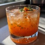 Close-up of a vibrant orange Kimchi Cocktail with Fermented Cabbage, ice, and a rosemary garnish.