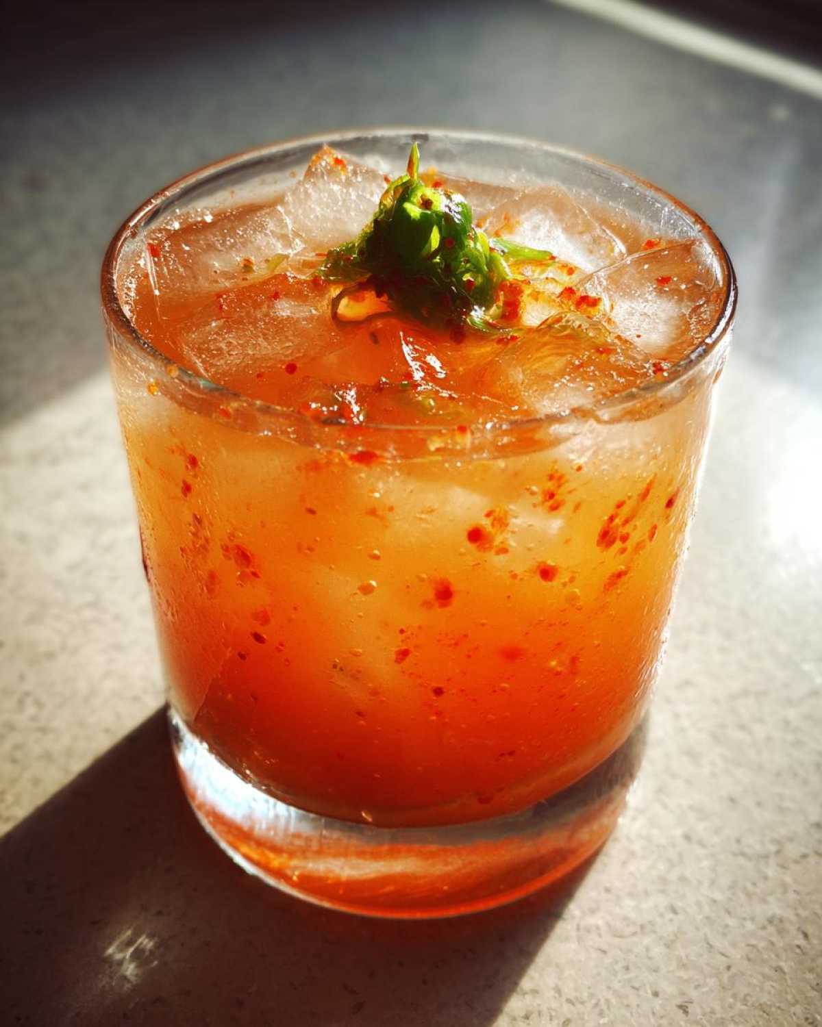 Close-up of a vibrant orange Kimchi Cocktail with Fermented Cabbage, served over ice and garnished with green chili.