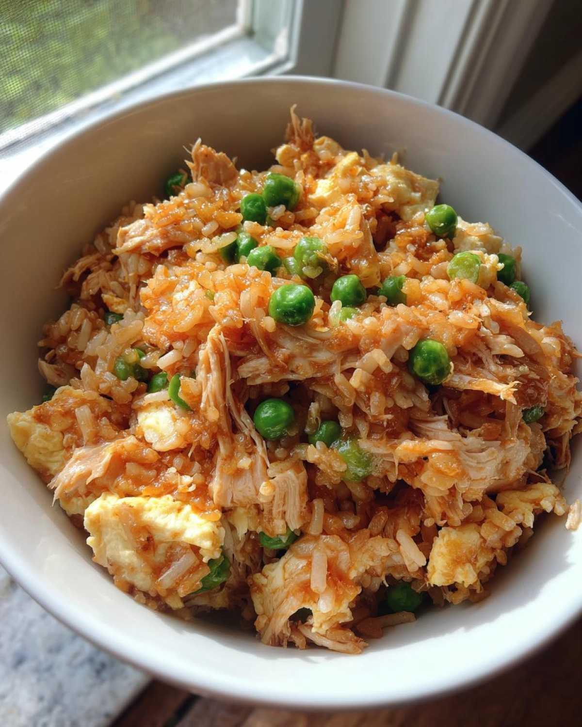 Close-up of a white bowl filled with Leftover Rotisserie Chicken Fried Rice, featuring shredded chicken, rice, scrambled egg, and bright green peas.