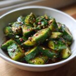 A white bowl filled with smashed, spicy Logan Moffit's Din Tai Fung Cucumber Salad, glistening with dark sauce and chili flakes.