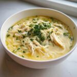 A close-up of a white bowl filled with rich Mediterranean Lemon Chicken Soup with Orzo, topped with shredded chicken and fresh dill.