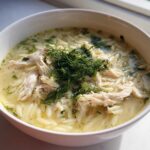 Close-up of a white bowl filled with Mediterranean Lemon Chicken Soup with Orzo, topped with shredded chicken and fresh dill.