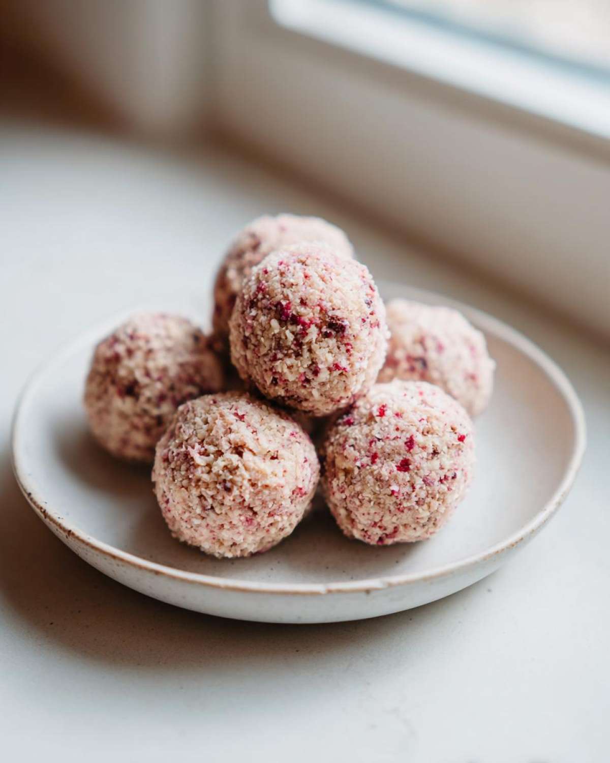 A stack of six No-Bake Strawberry Shortcake Protein Bites coated in coconut flakes, resting on a small ceramic plate.