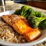 Close-up of a glazed salmon fillet topped with sesame seeds served over brown rice with roasted broccoli in an Orange Ginger Salmon Bowls preparation.