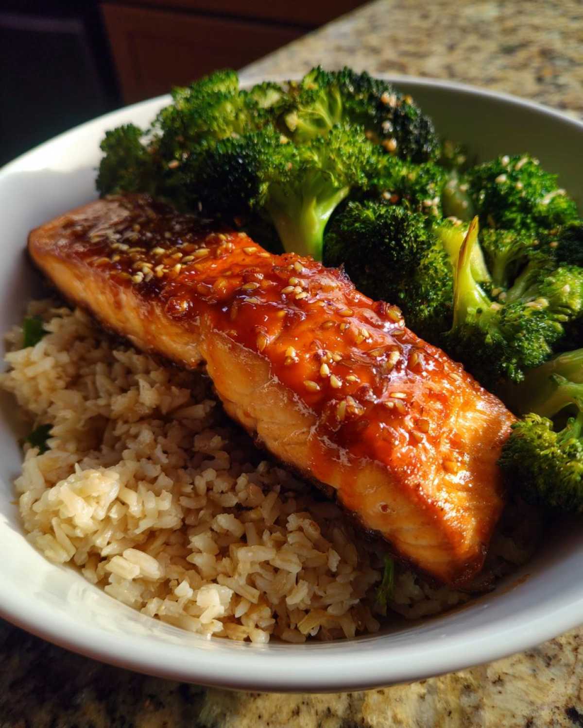 A piece of glazed salmon topped with sesame seeds served over brown rice with steamed broccoli in Orange Ginger Salmon Bowls.
