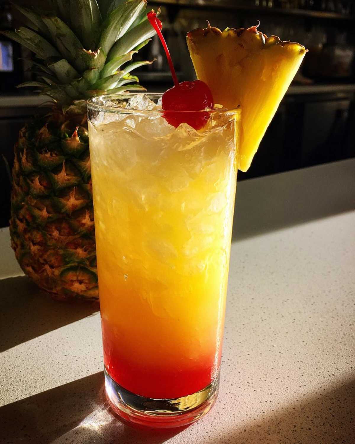 A tall glass of Piña Colada Sunrise Mocktail showing red, orange, and yellow layers, garnished with a pineapple wedge and cherry.