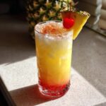 A layered Piña Colada Sunrise Mocktail with crushed ice, garnished with a pineapple wedge and a maraschino cherry.