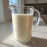 A glass pitcher filled with creamy Salted Maple Cashew Milk sitting on a bright white countertop.