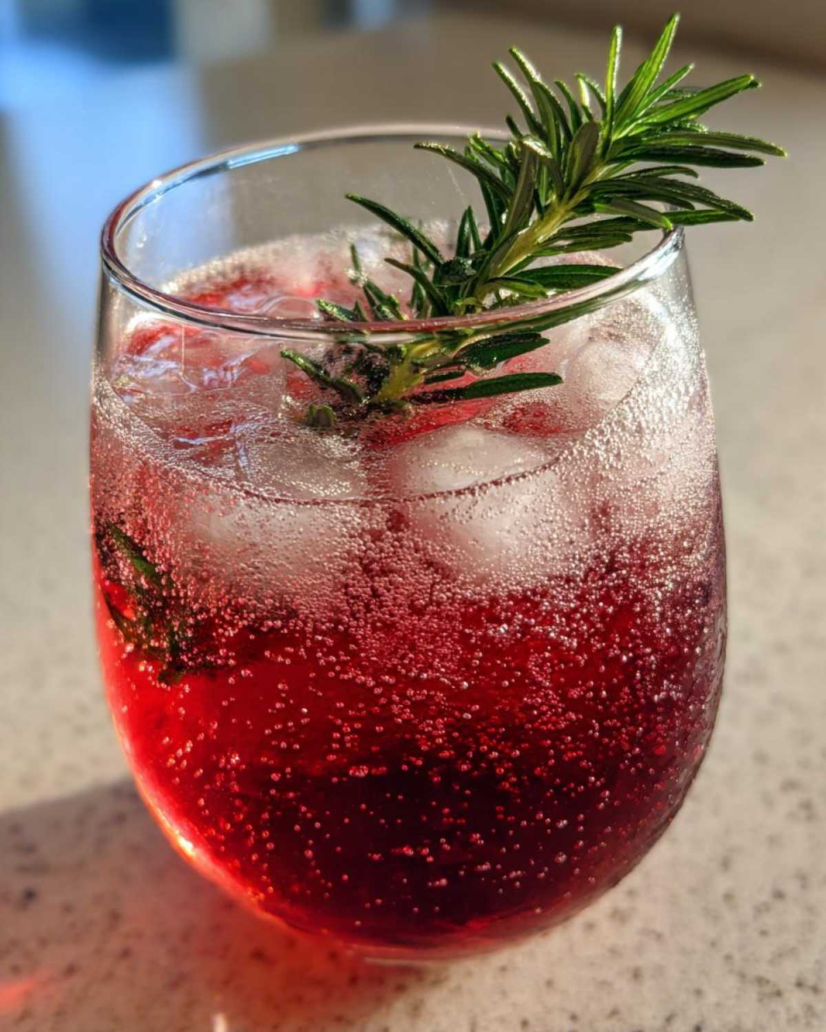Close-up of a fizzy, deep red Pomegranate Spritz with Rosemary garnish and ice cubes.