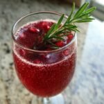 A close-up, top-down view of a fizzy, deep red Pomegranate Spritz with Rosemary garnish and floating pomegranate seeds.