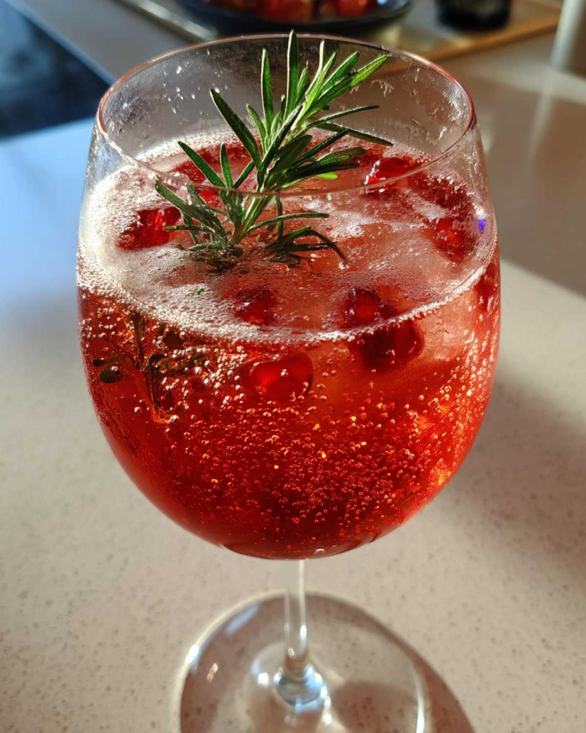 Close-up of a vibrant red Pomegranate Spritz with Rosemary garnish and floating pomegranate seeds.