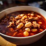 Close-up of a white bowl filled with rich, red Pozole Rojo with Pork and Hominy.