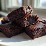 A stack of fudgy, dark chocolate Protein Brownie Bites with Collagen squares resting on a white plate near a window.
