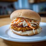 A close-up of a Roasted Cabbage Burger topped generously with spicy slaw, served on a white plate.