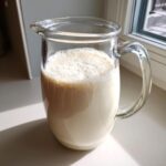 A clear glass pitcher filled with creamy Salted Maple Cashew Milk, showing a layer of froth on top, sitting by a sunlit window.