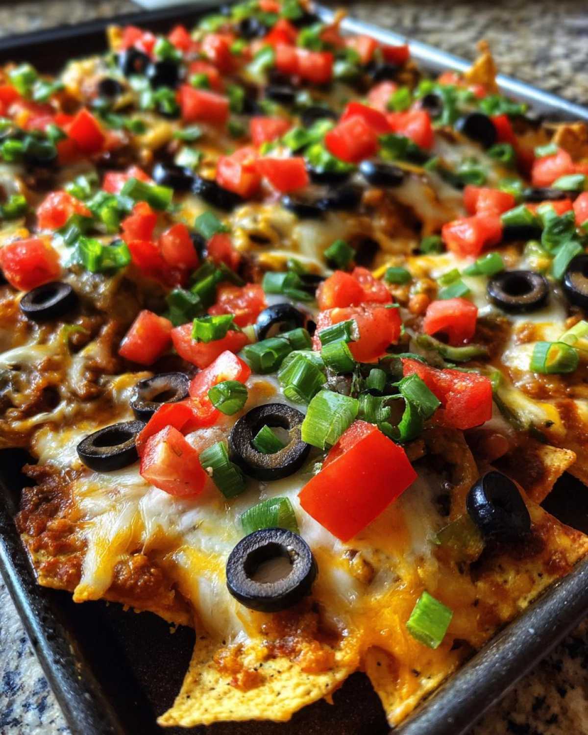 Close-up of freshly baked Sheet Pan Nachos with Refried Beans, topped with melted cheese, black olives, diced tomatoes, and green onions.