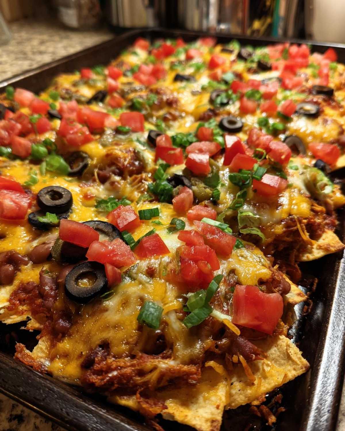 Close-up of fully loaded Sheet Pan Nachos with Refried Beans, melted cheese, tomatoes, olives, and green onions.