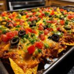 Close-up of fully loaded Sheet Pan Nachos with Refried Beans, melted cheese, olives, tomatoes, and green onions.