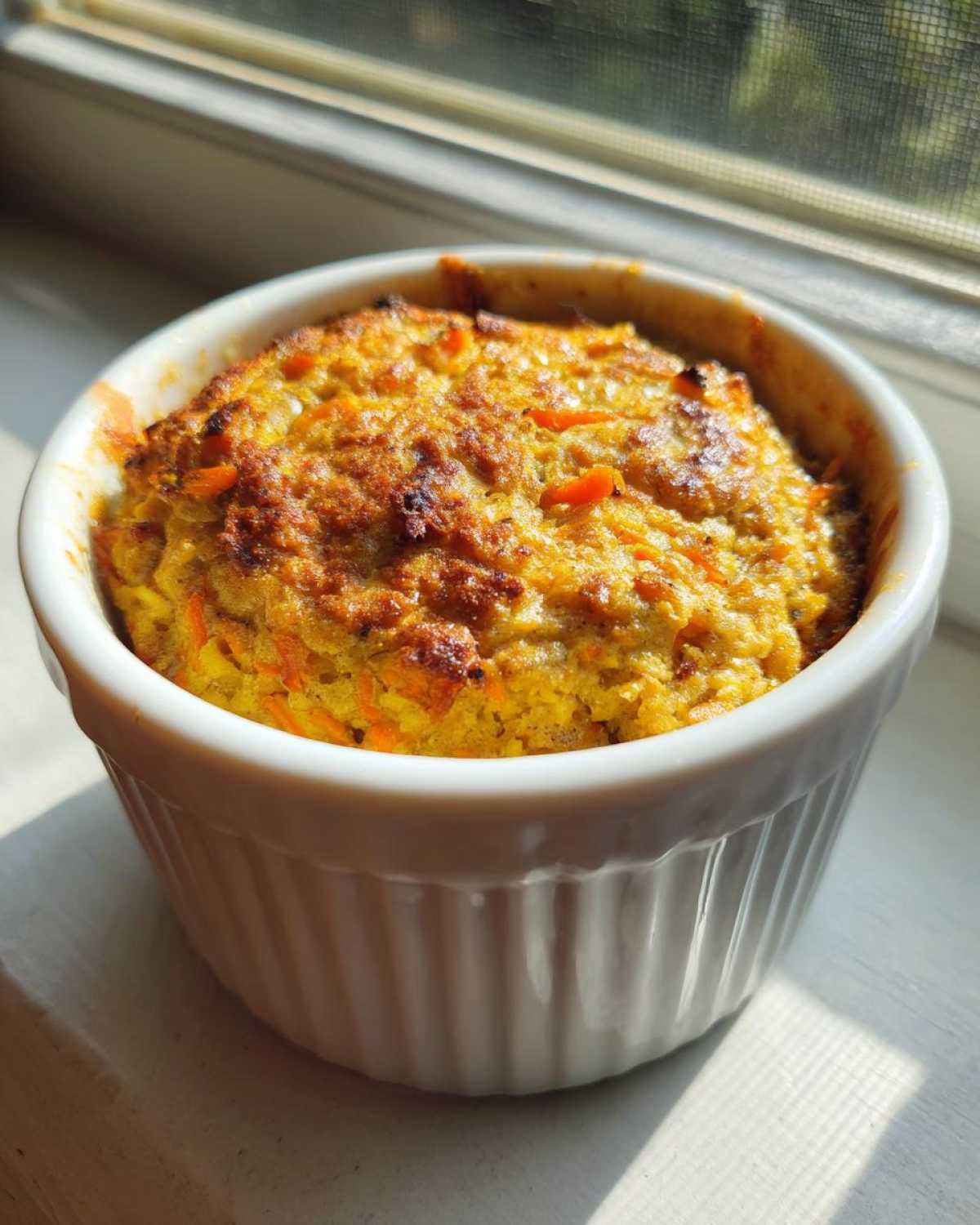 A golden-brown Single-Serving Carrot Cake Baked Oats baked in a white ramekin, sitting near a sunlit window.