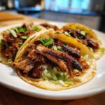 Close-up of four delicious Slow-Cooked Pork Carnitas Tacos filled with shredded meat, lettuce, and cilantro on a white plate.