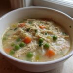 A close-up of a white bowl filled with creamy Slow Cooker Chicken Pot Pie Soup, featuring shredded chicken, carrots, and bright green peas.